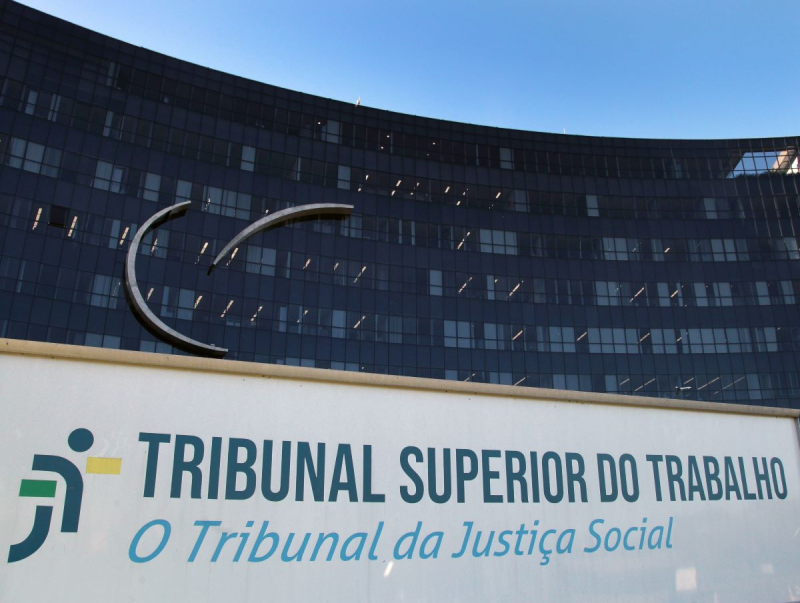 Fachada espelhada do TST no fundo com a placa na frente escrito: Tribunal Superior do Trabalho, o Tribunal da Justiça Social